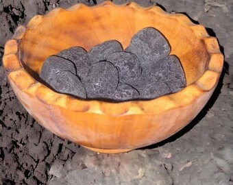 May include: A wooden bowl filled with smooth, black volcanic rocks. The bowl is sitting on a dark, rocky surface.