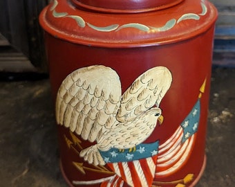 May include: Red metal canister with a painted design of a white eagle with outstretched wings holding an American flag. The eagle is perched on a bundle of arrows.