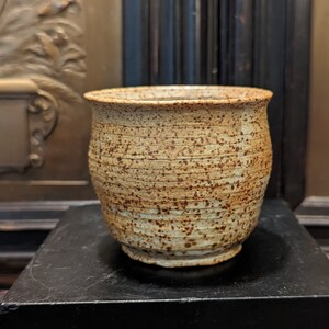 May include: A speckled brown and white ceramic mug with a rounded shape. The mug is sitting on a black surface.