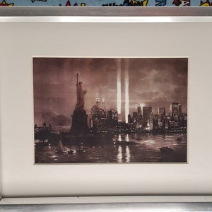 May include: A framed sepia-toned print of the New York City skyline with the Statue of Liberty in the foreground. The Twin Towers are visible in the background, with smoke rising from them.