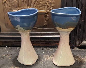 May include: Two ceramic cups with blue and white glaze. The cups have a wide, rounded bowl and a tall, slender stem. The cups are sitting on a dark surface.
