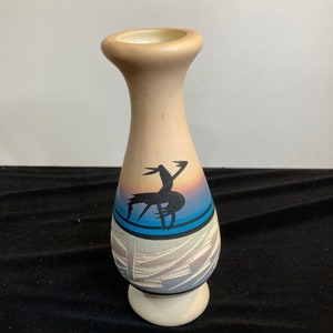 May include: A small, beige ceramic vase with a stylized Native American design. The vase has a blue, pink, and white design with a black silhouette of a bird.
