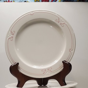 May include: A stack of ten white ceramic dinner plates with a delicate red floral design around the rim. The plates are stacked on a wooden stand.
