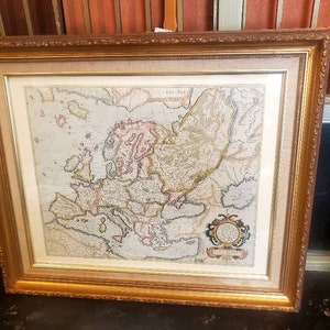 May include: A framed antique map of Europe with a decorative border and a title cartouche in the lower right corner. The map is printed on paper and has a cream-colored background.