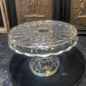 May include: A clear glass cake stand featuring a circular top with a raised grid pattern and a scalloped edge. The stand is supported by a matching pedestal base with a textured, repeating geometric design that extends from the center.