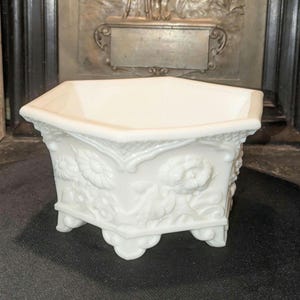 May include: A hexagonal white milk glass planter featuring embossed floral patterns on each side. The vessel has a wide, open top rim and is supported by four small, decorative feet at the base, showcasing intricate relief details throughout the exterior.