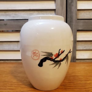 May include: White ceramic vase with a black and green floral design. The vase has a round body and a narrow opening.