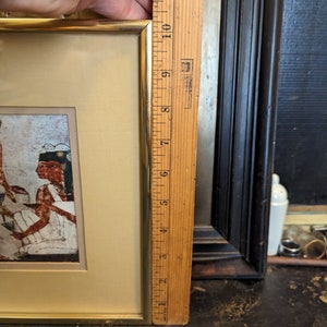 May include: A framed picture with a gold frame and a beige mount. The picture depicts a scene of people in ancient Egyptian clothing. The picture is 17.8 cm tall.