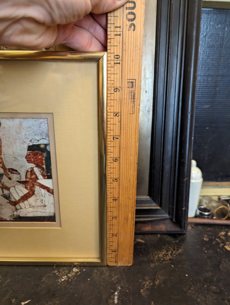 May include: A framed picture with a gold frame and a beige mount. The picture depicts a scene of people in ancient Egyptian clothing. The picture is 17.8 cm tall.