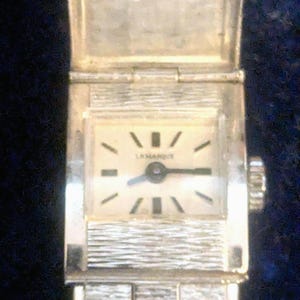 May include: A silver-toned wristwatch with a rectangular case and a textured metal link bracelet. The watch face is off-white with black hour markers and the text LAMARQUE printed in black. It features a hinged cover over the watch face.