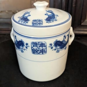 May include: White ceramic jar with a lid, adorned with blue fish and Chinese characters. The jar has a cylindrical shape with small handles and a knob on the lid. A decorative piece for the home.