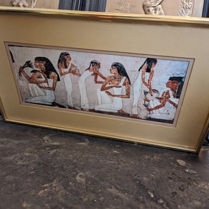 May include: A framed print of an ancient Egyptian scene showing seven women in white robes. The women are standing in a line, with the woman at the front holding a lotus flower. The print is set against a beige background.
