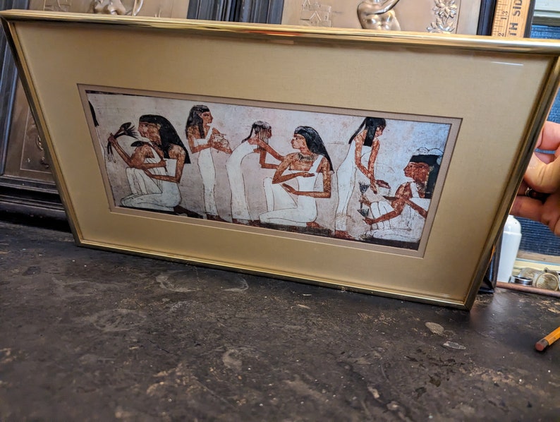 May include: A framed print of an ancient Egyptian scene showing seven women in white robes. The women are standing in a line, with the woman at the front holding a lotus flower. The print is set against a beige background.
