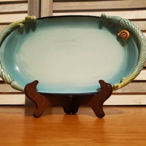 May include: A light blue oval serving dish with green and yellow leafy details and a small snail on one side. The dish is on a wooden stand.