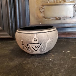 May include: A small, round, white ceramic bowl with black painted designs. The bowl has a geometric pattern and a stylized figure with a long tail.