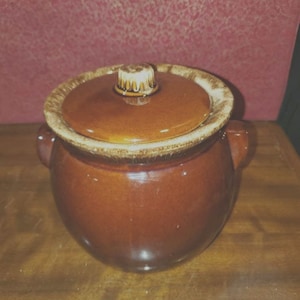 May include: A brown ceramic crock with a lid. The crock has a glazed finish and a rounded shape. The lid has a knob on top.