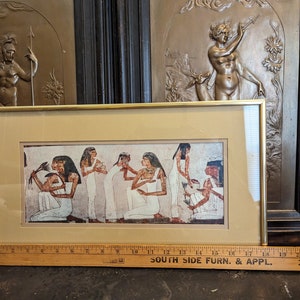 May include: A framed print depicting an ancient Egyptian scene featuring women in traditional clothing and jewellery. The print is set against a beige background and has a gold-coloured frame.