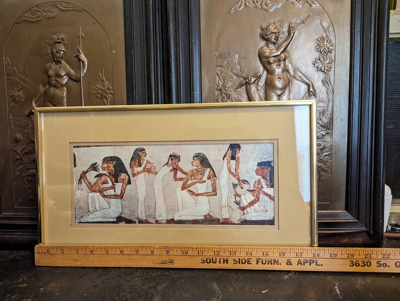 May include: A framed print depicting an ancient Egyptian scene featuring women in traditional clothing and jewellery. The print is set against a beige background and has a gold-coloured frame.