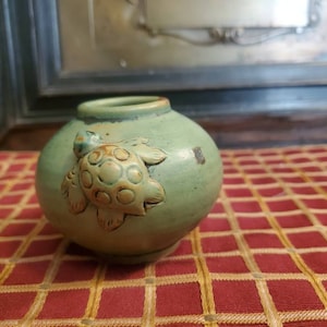 May include: A small, green ceramic vase with a raised, textured turtle design. The vase has a glazed finish and is perfect for displaying small flowers or plants.