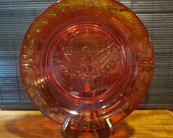 May include: A red glass plate with a raised design of an eagle with outstretched wings and a wreath of leaves. The plate has a gold rim and the words "In God We Trust" are etched around the edge.