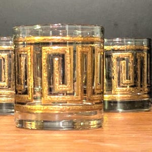May include: Set of six clear glass tumblers with a gold and black geometric design. The glasses have a rectangular pattern around the body and a gold rim. The glasses are cylindrical and sit on a wooden surface.