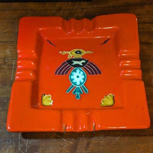 May include: Orange ceramic ashtray with a stylized bird design in shades of yellow, green, white, and brown. The bird has a large eye and a geometric pattern.