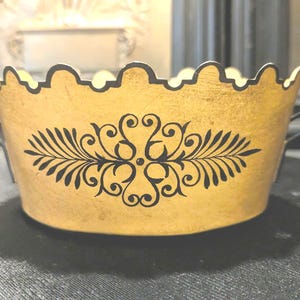 May include: A decorative, gold-colored metal container with a scalloped rim and two small handles. The container features a black, symmetrical floral design on the front. The container is likely used for decorative purposes.