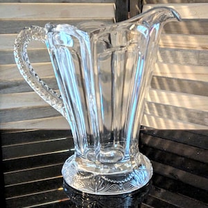 May include: A clear glass pitcher with a decorative, faceted design and a diamond-shaped handle. The pitcher has a wide mouth and a base with a detailed, etched pattern.