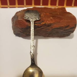 May include: Silver spoon with a detailed design of a small house on the handle. The spoon is resting on a piece of red rock.