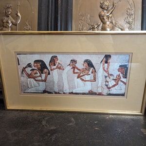 May include: A framed print of an ancient Egyptian wall painting depicting a group of women in white robes performing a ritual. The women are standing and sitting in a line, with their hands raised in a gesture of offering. The painting is set against a background of red and brown.
