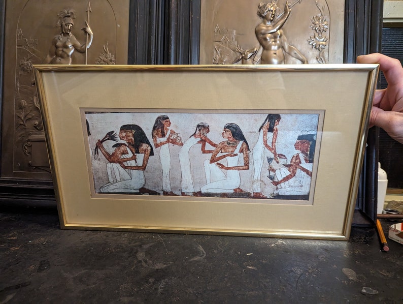 May include: A framed print of an ancient Egyptian wall painting depicting a group of women in white robes performing a ritual. The women are standing and sitting in a line, with their hands raised in a gesture of offering. The painting is set against a background of red and brown.