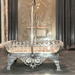 May include: An antique silver-plated serving dish with a clear glass insert. The dish has a square shape with scalloped edges and a decorative handle. The base features ornate floral and scrollwork details, adding to its vintage charm.