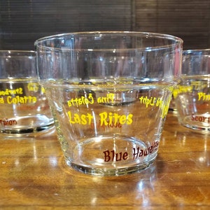 May include: Four clear glass tumblers with yellow and red lettering. The tumblers have different designs and text, including "Last Rites", "Blue Hawaiian", "Pina Colada", and "Scorpion".