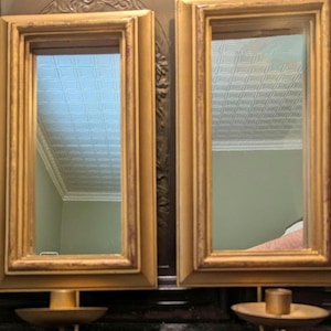 May include: Two gold-framed mirrors with ornate details, each reflecting a white ceiling with a patterned design. The mirrors are mounted on a dark surface.