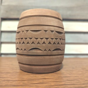 May include: A brown wooden cup with a carved geometric pattern. The cup is cylindrical and has a smooth, polished finish.