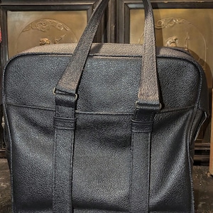 May include: Black briefcase with a textured surface and two top handles. The bag has a rectangular shape with a front pocket and metal hardware. The briefcase is designed for carrying documents and other items.