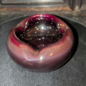 May include: A round, deep glass ashtray featuring a smooth, polished surface with a rich, dark burgundy and plum color gradient. The thick-walled vessel has three distinct indentations along the rim and contains small, scattered air bubbles.