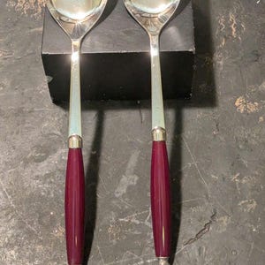 May include: A pair of silver serving spoons with a slotted spoon and a solid spoon. The handles are a deep burgundy color with silver accents. The spoons are displayed on a dark surface.