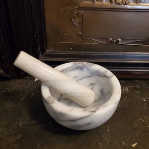 Puede incluir: Un mortero y un pilón de mármol blanco con vetas grises. El mortero es redondo y el pilón es cilíndrico.