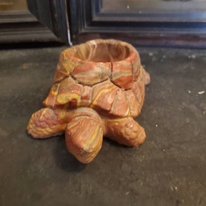 May include: A small, ceramic turtle planter with a red, brown, and yellow striped pattern. The planter has a round opening on top for planting.
