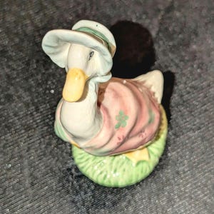 May include: A ceramic duck figurine wearing a light blue hat with a green band and a pink dress with green shamrock accents. The duck is perched on a green grassy base, with a yellow beak.