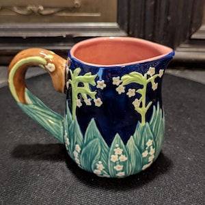May include: A ceramic pitcher with a dark blue exterior, decorated with green leaves and white flowers. The handle is brown with green accents. The interior is pink. The pitcher has a spout for easy pouring.