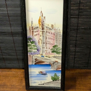 May include: A framed tile art piece depicting a European cityscape with a bridge, river, and buildings. The artwork features a boat on the water and trees. The frame is black, and the art is in a vertical orientation.