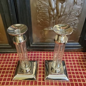 May include: A pair of silver candle holders with clear glass cylinders. The candle holders have a square base and a round top. The candle holders are on a red and white checkered surface.