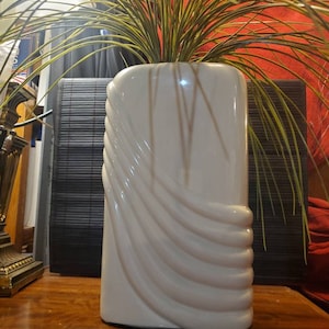 May include: A white ceramic vase with a wavy, textured design. The vase is filled with green artificial grass.