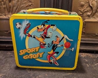 May include: A yellow metal lunchbox featuring Goofy and a cartoon character playing basketball and soccer. The lunchbox has the words "Sport Goofy" printed on the front.