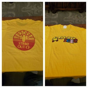 May include: Yellow t-shirt with a red and white bowling pin graphic on the back and a Pac-Man graphic on the front. The back graphic says "Strike Out CF".