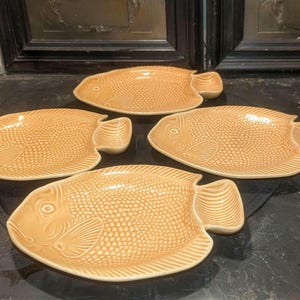 May include: Four ceramic fish-shaped serving dishes. The dishes are a warm, light orange color with a textured scale pattern. Each dish has a fin-shaped handle on one side. The dishes are arranged on a dark surface.