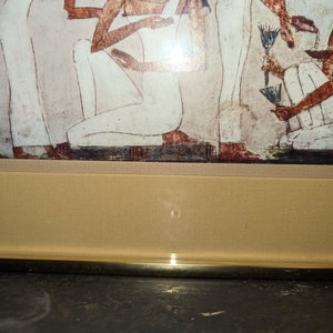 May include: A framed print of an ancient Egyptian wall painting depicting a group of people in white robes. The painting is in a gold frame with a beige mount.