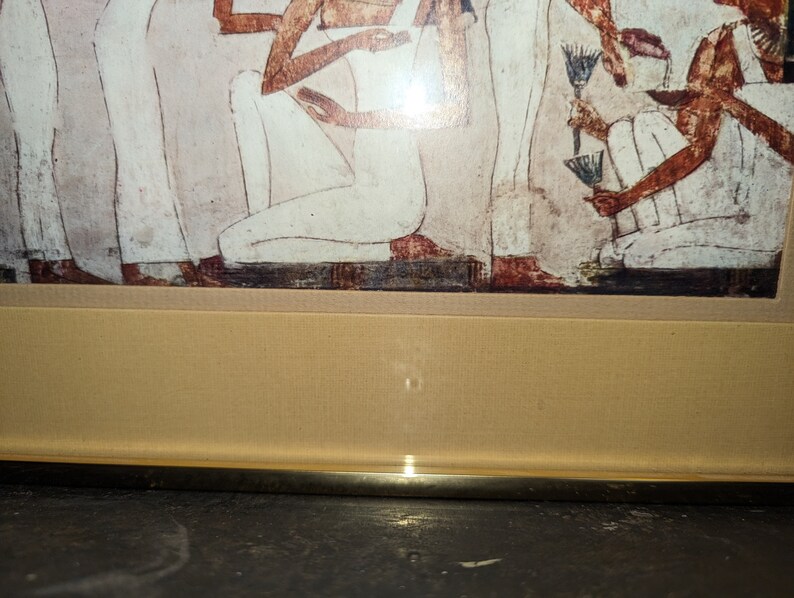 May include: A framed print of an ancient Egyptian wall painting depicting a group of people in white robes. The painting is in a gold frame with a beige mount.